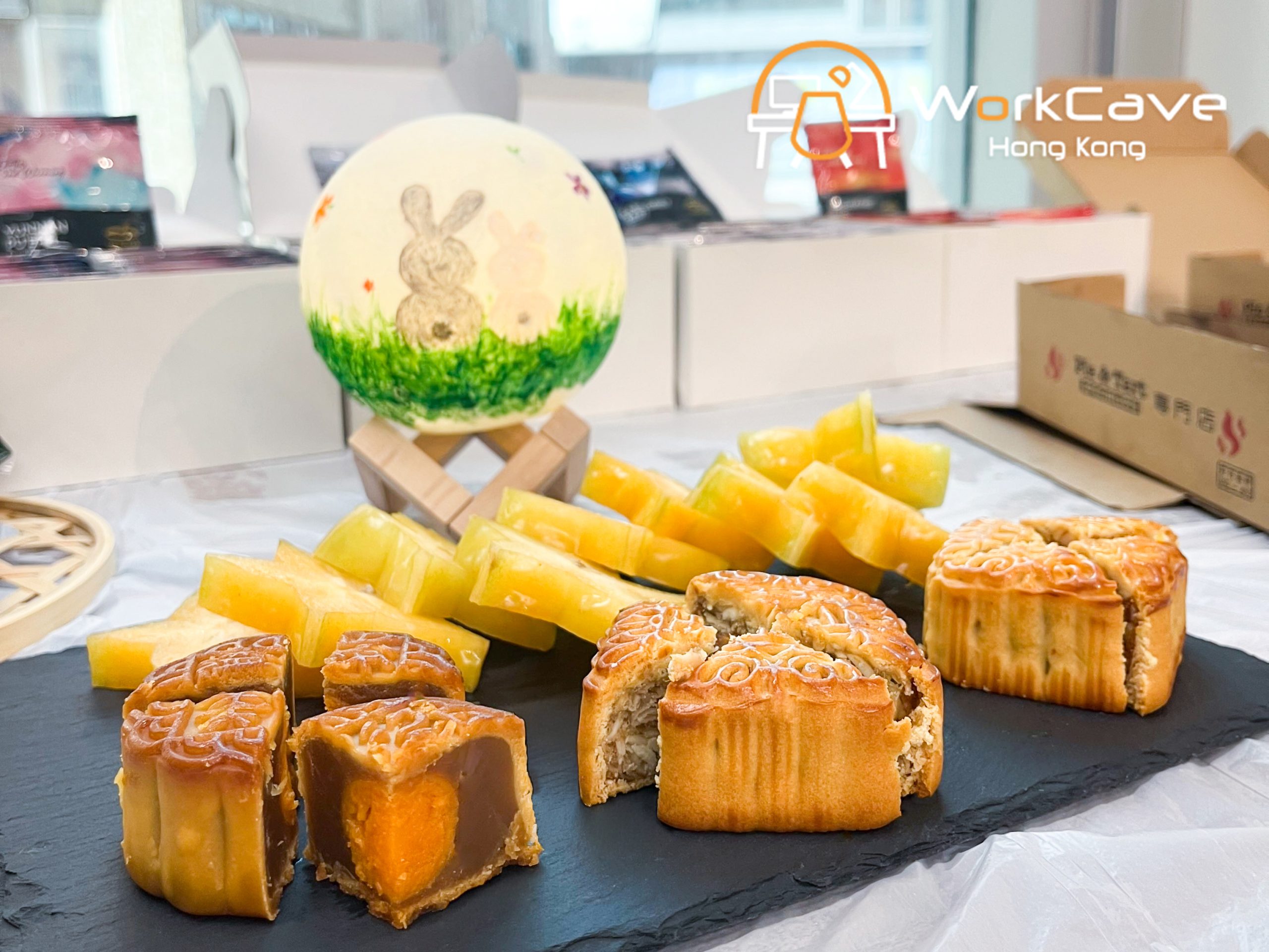 mooncakes and star fruits on a plate, behind is a moon lamp painted the back of one grey and one pink rabbit which are sitting on the grass
